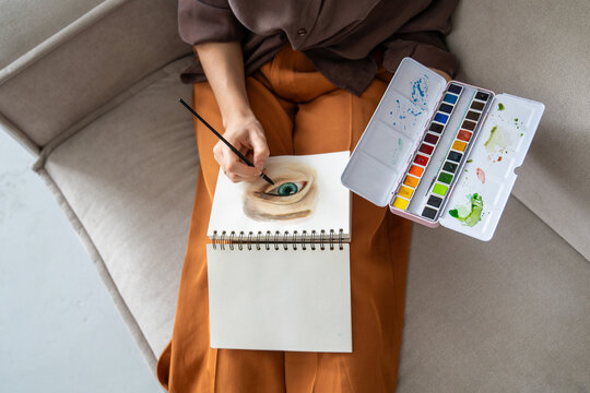 Woman enjoying peaceful time at home and art hobby, sitting on cozy couch painting picture with watercolor, female artist working on new drawing with dedication, upgrading skills, getting inspiration.