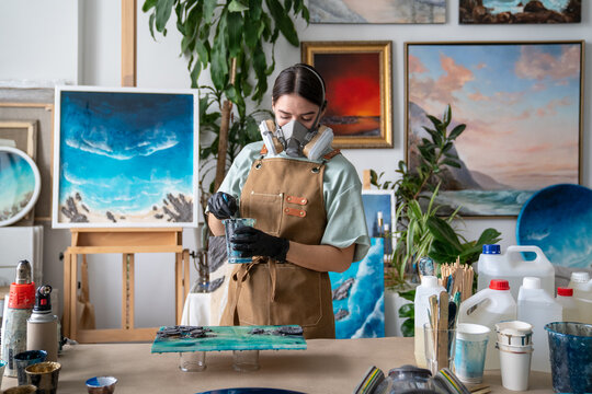 Woman in respirator mixing varnish and epoxy in cup at workshop table, creative female artist working on liquid art, using supplies and equipment for epoxy resin craft, working process in gallery.