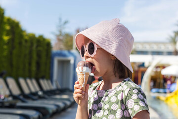 Little girl wearing pink bucket hat and stylish sunglasses eating melting ice cream cone near poolside loungers in sunny resort, summer vacation lifestyle with playful mood, childhood joy, copy space © Evgeniia Primavera