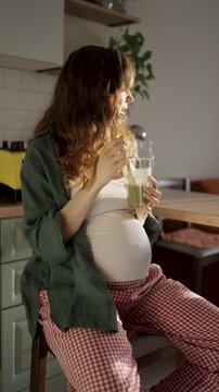 Pregnant woman enjoying a glass of fresh green smoothie while sitting in her sunlit kitchen, gently caressing her belly and maintaining a healthy diet for her baby's well-being