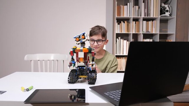 Young boy in glasses studies a colorful block built robot at a table with laptop and tablet, illustrating hands on stem learning, curiosity, coding and creative problem solving