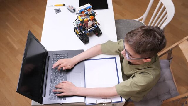 Young boy focused on robotics and coding at a desk, building a colorful robot with a laptop for a stem project, exploring programming, electronics and problem solving