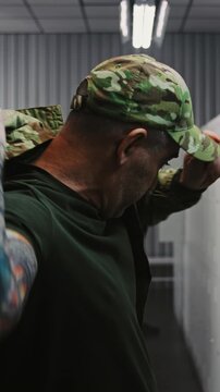 Soldier With Tattooed Arms Puts on Camouflage OCP Uniform Jacket and Cap by Locker, Getting Ready For Tactical Mission While Colleagues Interact in Background. Military Base Preparation. Vertical Shot