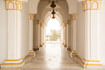 Fototapeta premium Mosque riwaq arcade with marble columns and pointed arches featuring gold capitals and hanging lanterns forming symmetrical passage. Islamic architecture, structural element, exterior space