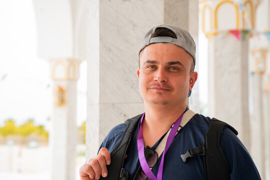 Caucasian male tourist standing in mosque courtyard with marble columns wearing casual clothes backpack and cap looking at camera. Travel, tourism, people, architecture