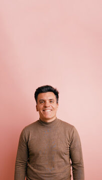 Cheerful middle aged man in brown turtleneck smiling warmly at camera against a soft pink studio background, confident and friendly