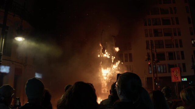 Large group of silhouetted people on city street at night watching tall burning falla structure collapse in orange flame, one person filming on smartphone