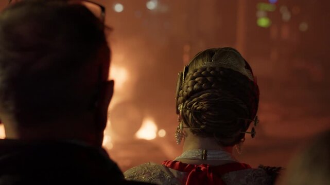 Back view of young woman in elaborate traditional fallera costume with ornate braided hairstyle, jeweled pin, pearl earring, embroidered golden dress with red sash, watching bonfire