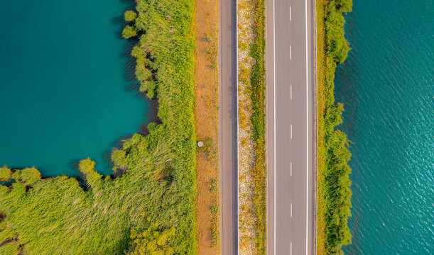 Top down aerial view of a road crossing a turquoise mountain lake with lush green vegetation. Minimalist travel landscape captured from above.
