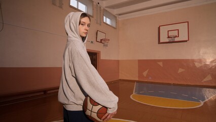 Young woman in casual sportswear stands confidently with a basketball in an old school gym, blending urban style and athletic confidence for youth sports and lifestyle imagery © Aliaksandr