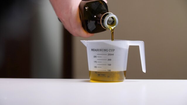 Person pouring healthy olive oil from a dark bottle into a marked clear measuring cup, preparing ingredients for cooking or baking, showing precise measurement in a kitchen setting