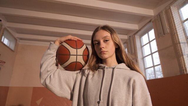 Teenager standing in an old school gym, looking at the camera while holding a basketball on her shoulder, symbolizing youth sports, practice, and a competitive spirit