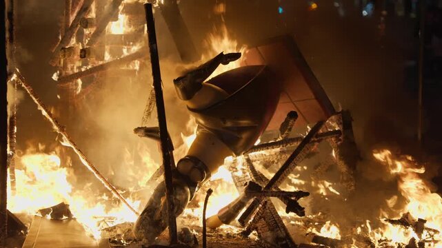 Close up of colorful painted ninot figure, stylized cartoon character in yellow, black and red, engulfed in flame and surrounded by burning debris during crema bonfire