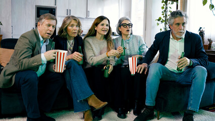 Fototapeta premium Group of senior friends sitting on sofa, eating popcorn and drinking beer while watching an exciting film at home