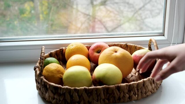 Woven basket on windowsill holds grapefruit, apples, orange, lime. Girl touches and inspects fruits. Early spring outdoors, reminder to get enough vitamins. 