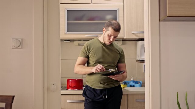 A man stands in his home kitchen, tasting food from a plate with a fork. He appears unhappy and dissatisfied with the taste of the meal, possibly finding it bland or unappetizing.