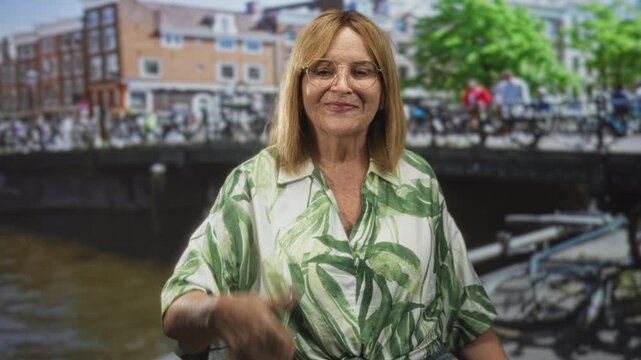 Woman with glasses gives thumbs up showing bare thumb on street by canal and bridge, wearing green leaf print blouse; optimism travel memory.