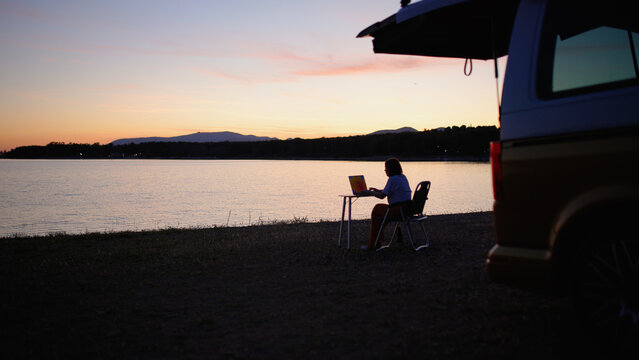 Woman using laptop at a portable desk on the shore, a camper van nearby, enjoying a peaceful remote work setting at sunset