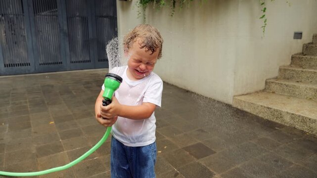Toddler boy plays with garden hose on hot summer day. He first sprays water to side, then turns hand sprinkler toward his own face, enjoying cool shower. Already soaked, its clear hes repeated joke