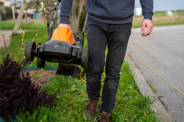Obraz premium Person carrying electric lawn mower beside lawn and roadside, showing portable garden equipment after mowing in outdoor daylight.