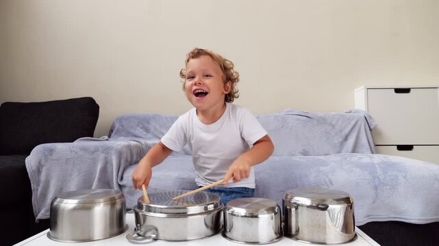 Cheerful young child has fun using kitchen pots and wooden sticks as makeshift drums, joyfully creating loud rhythmic sounds. Though not playing real music, young boy expresses creativity and energy