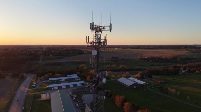 A cinematic 360-degree aerial drone orbit around a 5G cellular tower at golden hour, revealing rural farmland, agricultural buildings, and a vast horizon during a vibrant sunset
