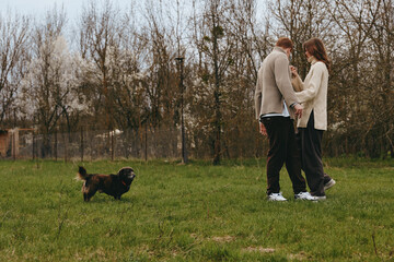 Naklejka premium Couple walking with a dog in a park, casual outfits, candid lifestyle moment, outdoor activity, gentle breeze, greenery and bare trees in the background, communicates bonding, happiness, and everyday