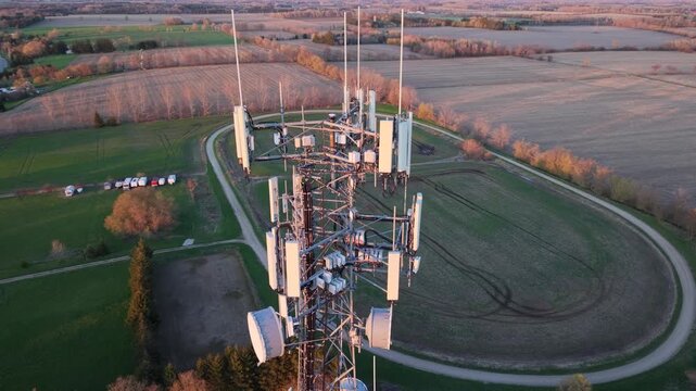 A cinematic aerial drone orbit around a lattice telecommunications tower during a vibrant golden hour, showcasing multiple antenna arrays and microwave dishes against a vast rural horizon