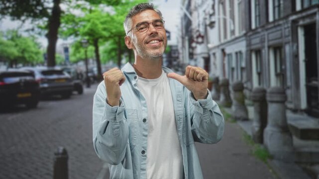 Middle aged man with thumbs pointing to chest on a cobblestone street beside row buildings, smiling and wearing glasses; confidence success.