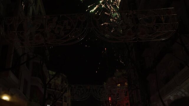 Colorful firework exploding through ornate Fallas street arch adorned with heart motif between residential building facade in Valencia, bare tree in foreground at night