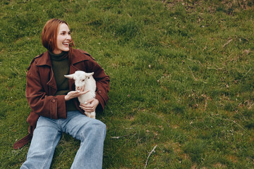 Naklejka premium Smiling woman sits on grass holding a kid goat in a park, wearing casual fashion. A warm lifestyle moment that celebrates human animal companionship outdoors, simple rural joy, and a relaxed sense of