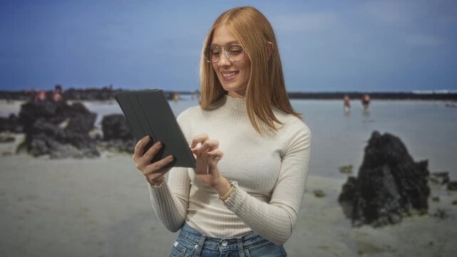 Woman tapping tablet with finger at sandy volcanic beach near shallow tide pools and rocks, smiling and wearing glasses; serenity focus.