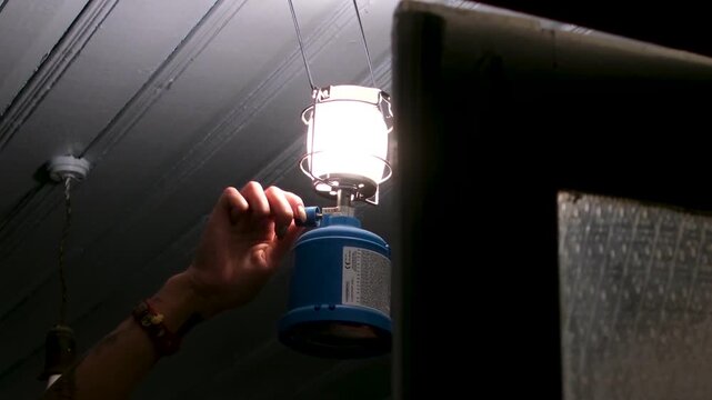 during a power outage, a woman carefully lights a kerosene lamp. The small flame begins to glow, softly illuminating the simple interior and creating a warm, calm nighttime village atmosphere. 