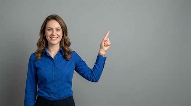 Happy young professional business woman wearing blue shirt looking at camera pointing finger up at copy space showing upward presenting advertising standing isolated at gray background vertical.