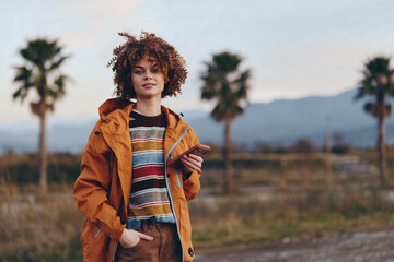 Fototapeta premium Woman lifestyle portrait outdoors, curly hair, orange jacket, striped sweater, standing on a rural road, palm trees and mountains in sunset light, relaxed travel mood