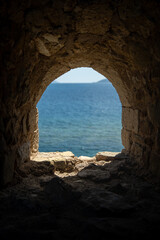 Fototapeta premium Ancient stone arch window of a medieval castle overlooking the blue Mediterranean Sea in Peloponnese, Greece. Historic frame-within-frame travel photography.