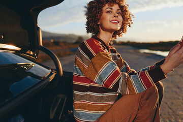 Fototapeta premium Woman lifestyle smiling at sunset while driving a car on an open road, hands on steering wheel, wearing a striped sweater, warm golden light, outdoor travel moment