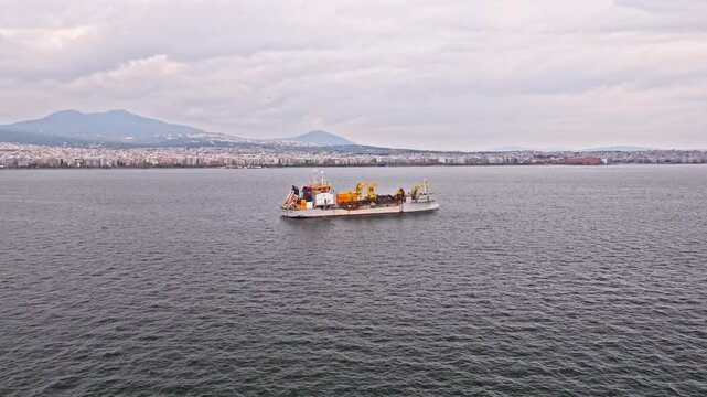 High-quality aerial footage of a dredger support vessel, also known as a subsea cable ship, operating in a busy industrial port. The vessel is designed for underwater cable installation, maintenance, 