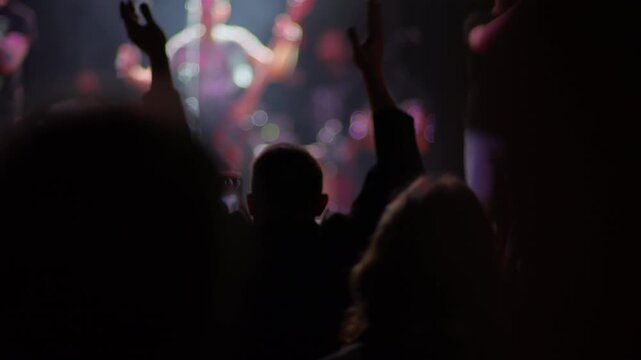 Fan claps raised hands at live concert, blurred performer and warm stage light visible behind, purple and warm bokeh fills background