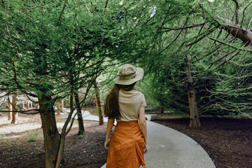 Fototapeta premium woman walking path trees hat back park skirt in green canopy, rear view of a traveler on a curved walkway surrounded by foliage for nature stroll and summer leisure