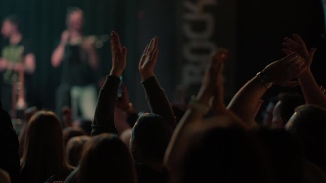 View from behind of an energetic crowd clapping hands at a live rock concert. People cheer for the band on a dimly lit stage with atmospheric lighting. A celebration of live music and nightlife
