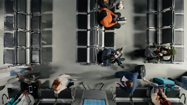 Travelers in airport terminal walking through people using devices on chairs. Passengers with trolleys navigating air transportation hub next to people waiting for plane, top down view shot