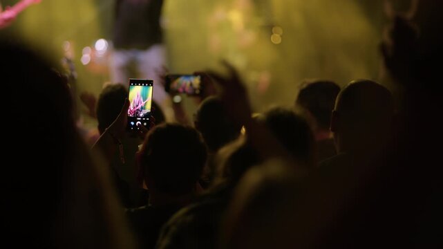 Concert fan holds phone in one hand and a cup in the other, neon strip light visible above, teal blue atmosphere fills background