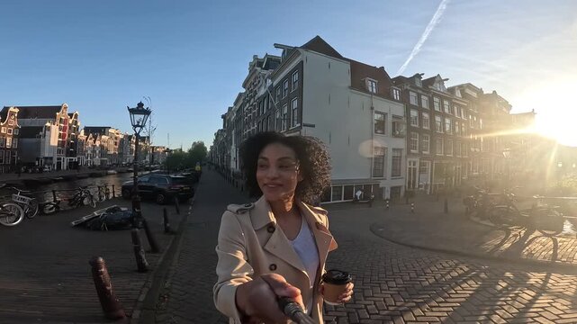 Influencer woman vlogging with selfie stick walking on a bridge in amsterdam during a sunny day and holding a coffee cup