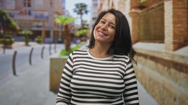 Woman smiling in a striped shirt, facing camera on a sunlit street with building wall and planters in view; joy warmth confidence.