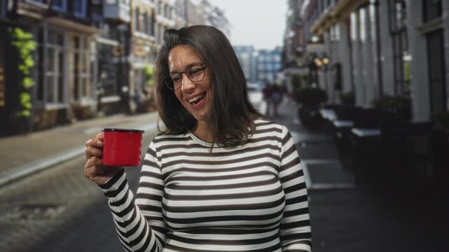 Woman holding a red enamel mug, smiling and tilting it toward camera while standing on a narrow cobblestone street outside a sidewalk cafe; contentment leisure.