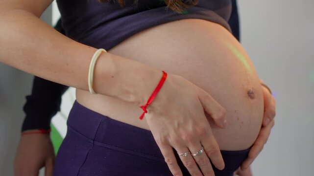 Profile close view of large pregnant belly, woman rests arm across lower abdomen, red thread bracelet visible on wrist, blurred plant visible in background
