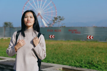 Fototapeta premium Woman with backpack in outdoor park beside a ferris wheel, casual attire enjoying sunny day and urban scenery