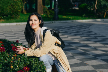 Fototapeta premium Woman kneeling beside flowers in a park, outdoor scene with backpack, casual attire, gentle smile, sunlight among greenery, calm nature moment suitable for stock photography, woman enjoying serene