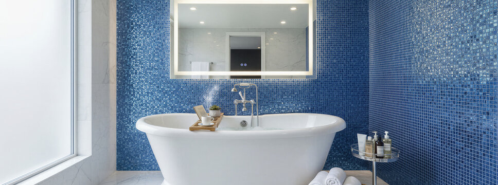 Modern bathroom interior with white freestanding bathtub against blue mosaic tile wall. Luxury home renovation and finishing work concept. Banner with copy space